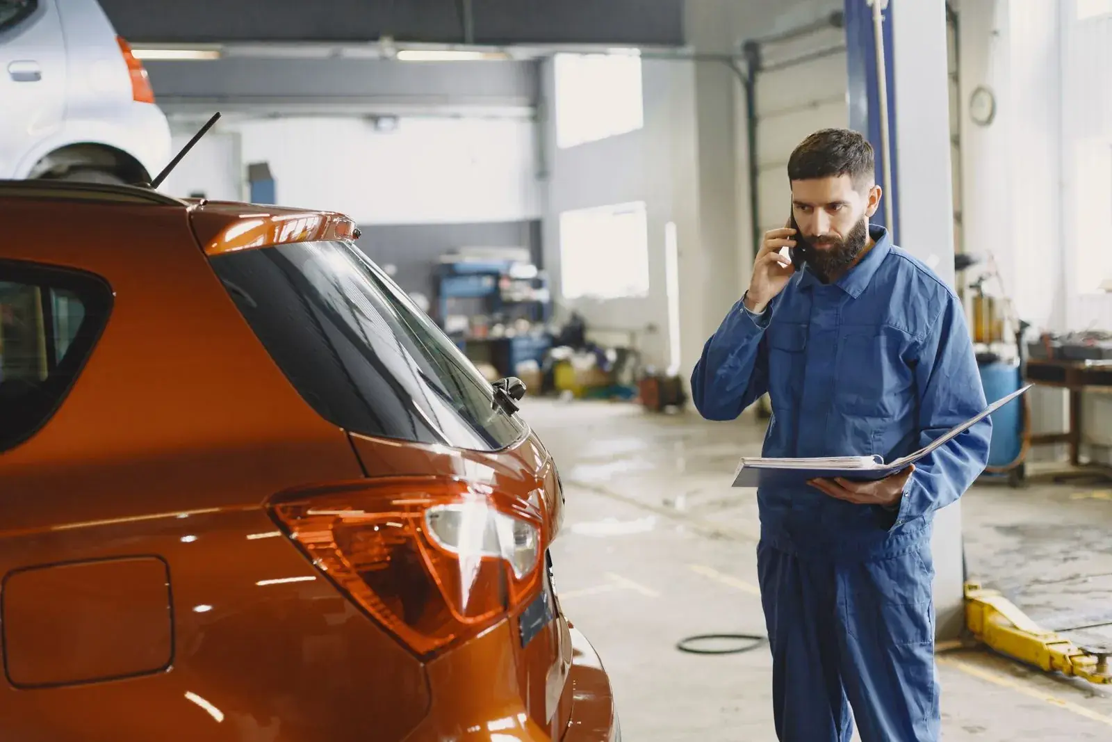 Un mécanicien au téléphone, un classeur ouvert à la main, près d'une voiture dans son garage