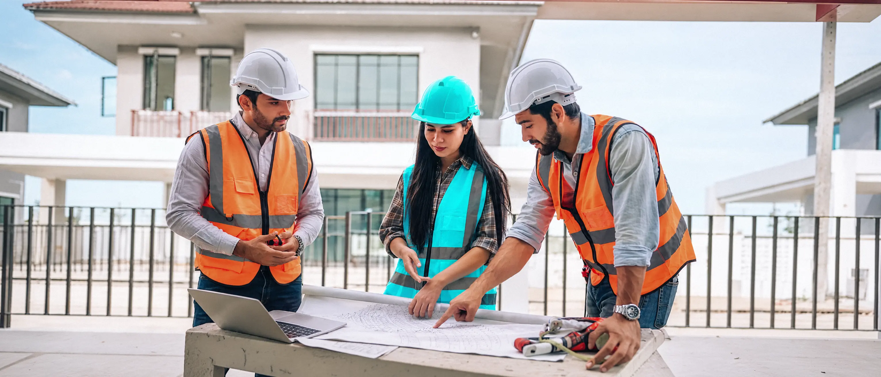 Trois ingénieurs en casque et gilet étudient un plan de construction avec un ordinateur portable sur le chantier